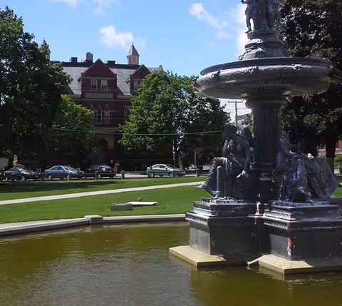 A decorative fountain in a shallow pond on a grassy lawn with a large brick building and parked cars in the background.