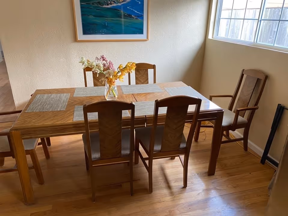 A wooden dining table with six matching chairs arranged around it. The table has six placemats and a vase with colorful flowers in the center. A large window on the right side lets in natural light, and a framed picture hangs on the wall above the table.
