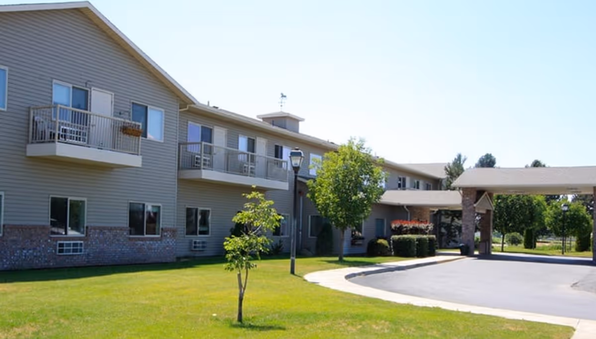 Exterior view of a two-story senior living facility building with balconies, windows, and a covered entrance driveway. The building is surrounded by a well-maintained lawn with small trees and shrubs under a clear blue sky.