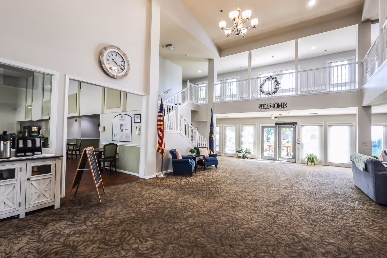 Spacious and well-lit senior living facility lobby with high ceilings, a large clock on the wall, a 'WELCOME' sign above the entrance, seating areas with blue chairs and a sofa, an American flag, and a coffee station on the left side.