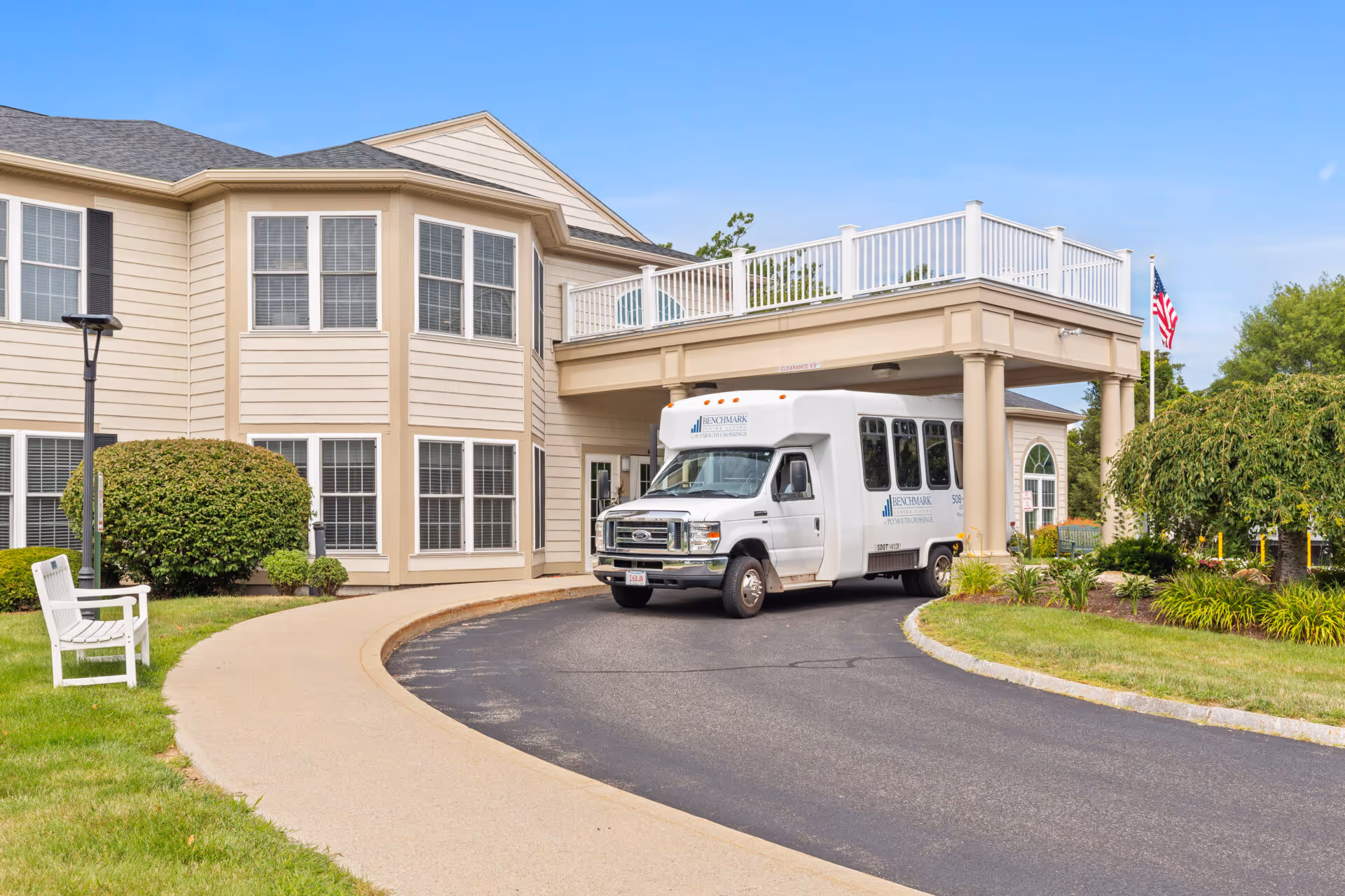 Exterior view of Benchmark Senior Living at Plymouth Crossings showing a beige two-story building with multiple windows, a covered entrance with columns, a white shuttle bus parked under the entrance canopy, green bushes, a white bench on the lawn, and an American flag in the background under a clear blue sky.