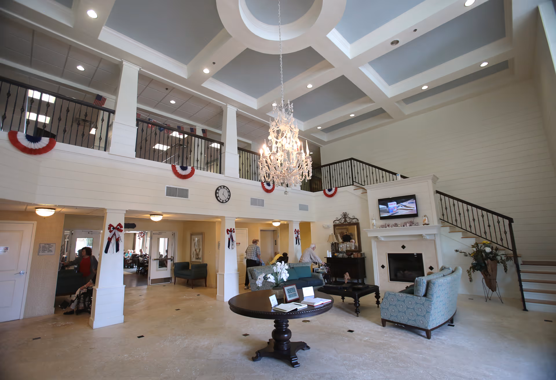 Spacious senior living facility common area with high coffered ceiling and chandelier. The room features a fireplace with a TV mounted above, comfortable seating including sofas and armchairs, a round table with decorative items, and a staircase leading to an upper balcony. Red, white, and blue patriotic decorations adorn the walls and railings. Several seniors are seen in the background near the entrance to another room.