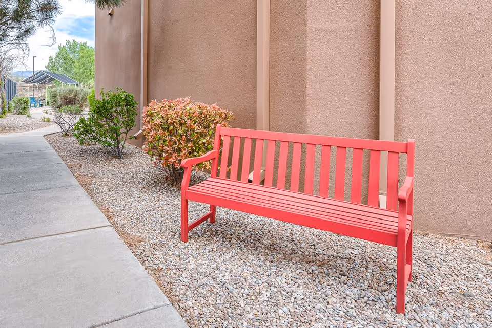 A red wooden bench on gravel beside a stucco building with shrubs and a concrete walkway.