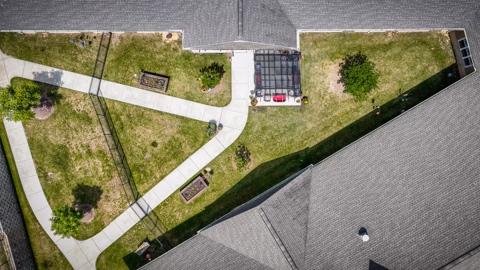 Aerial view of an outdoor garden area at Chatham Ridge Assisted Living, featuring intersecting concrete walkways, green grass, several small trees, and raised garden beds. The roof of the building surrounds the garden area, and there is a screened patio with outdoor furniture visible near the center top of the image.