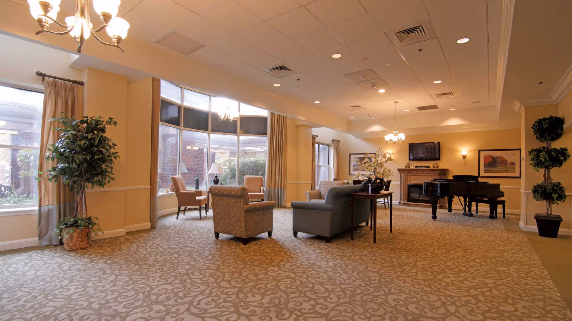A spacious and well-lit living room area with large windows allowing natural light to fill the space. The room features comfortable seating including armchairs and sofas arranged around a small table. There is a grand piano near a fireplace with a mounted TV above it. The walls are painted a warm beige color, and the floor is covered with a patterned carpet. Potted plants and framed artwork add decorative touches to the room.