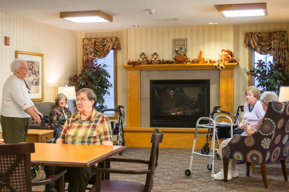 A cozy senior living common area with several elderly women sitting and standing around tables and chairs. The room features a fireplace decorated with autumn-themed ornaments, patterned curtains, potted plants, and warm lighting. Some women use walkers and are engaged in conversation or sitting quietly.