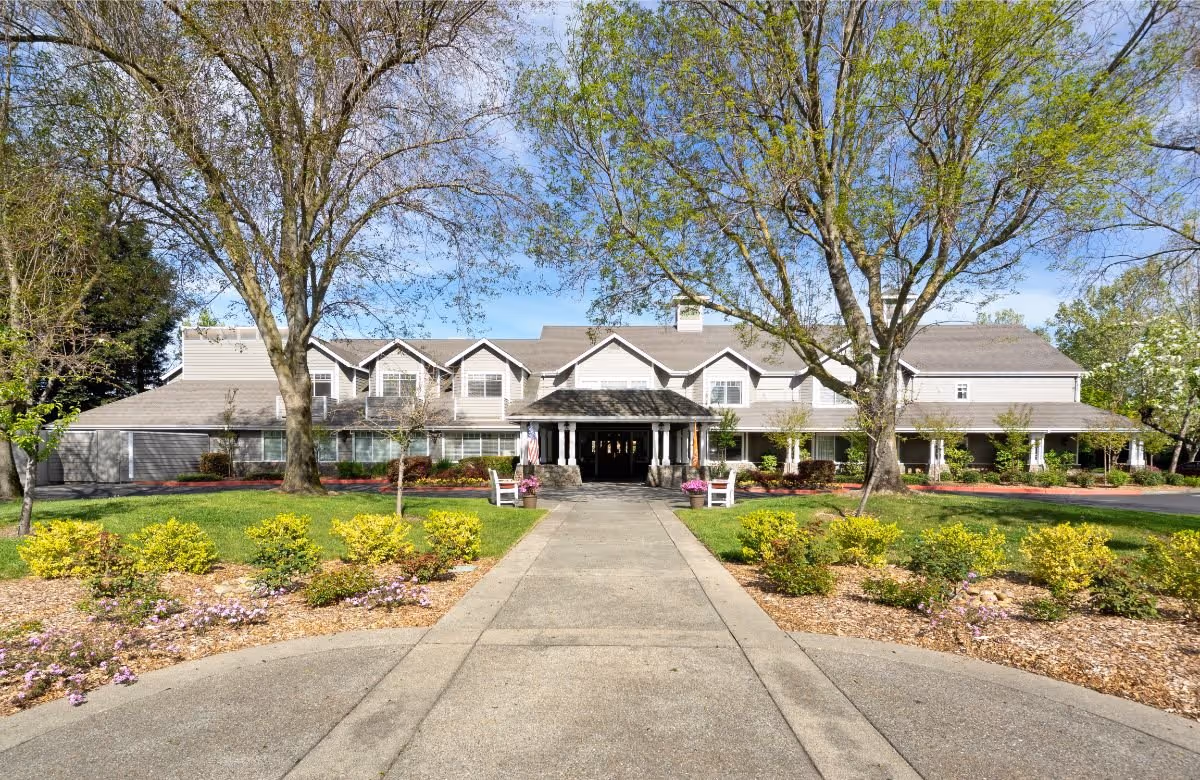 Front exterior view of Windsong of Sonoma Senior Living facility with a paved walkway leading to the entrance, surrounded by green lawns, trees, and landscaped flower beds under a clear blue sky.