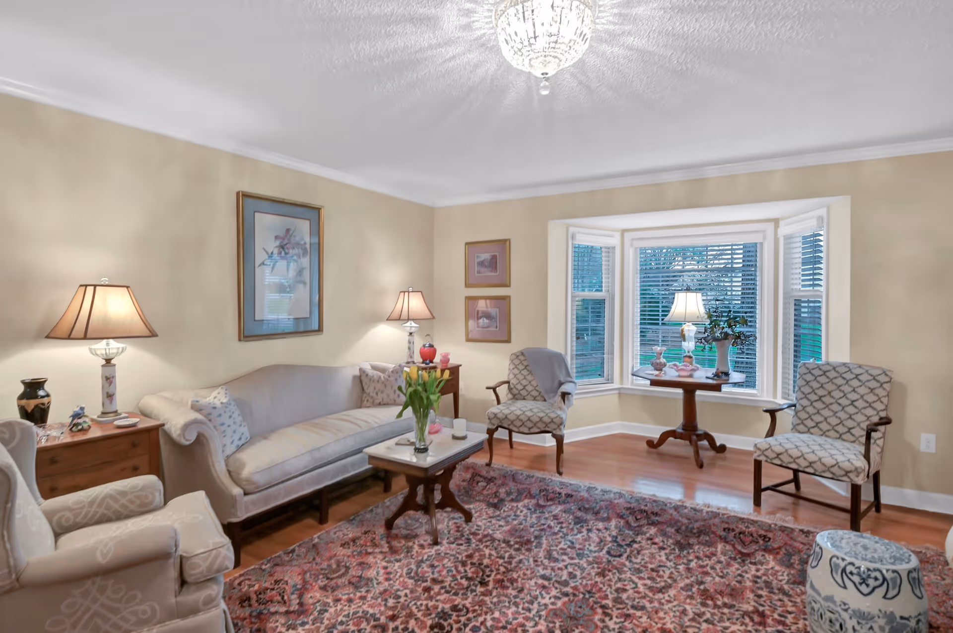 A cozy living room with beige walls and wooden flooring. The room features a light beige sofa with patterned cushions, two upholstered armchairs with a geometric pattern, a wooden coffee table with a vase of yellow tulips, and a side table with a lamp and decorative items. A large bay window with white blinds allows natural light to enter, and a small table with a lamp and plants is placed in front of the window. A patterned area rug covers the floor, and a crystal chandelier hangs from the ceiling.