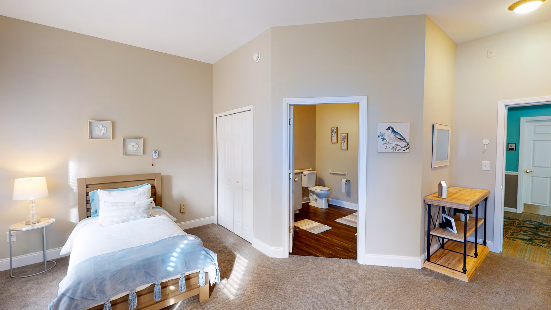 A bright and clean bedroom with a single bed made with white and light blue bedding. There is a small round side table with a lamp on the left side of the bed. The room has beige walls and carpeted floor. A closet with white double doors is visible next to an open doorway leading to a bathroom with a toilet and wooden floor. On the right side, there is a small wooden shelf unit with decorative items and a wall painting of a bird. Another doorway leads to a hallway with teal-colored walls.