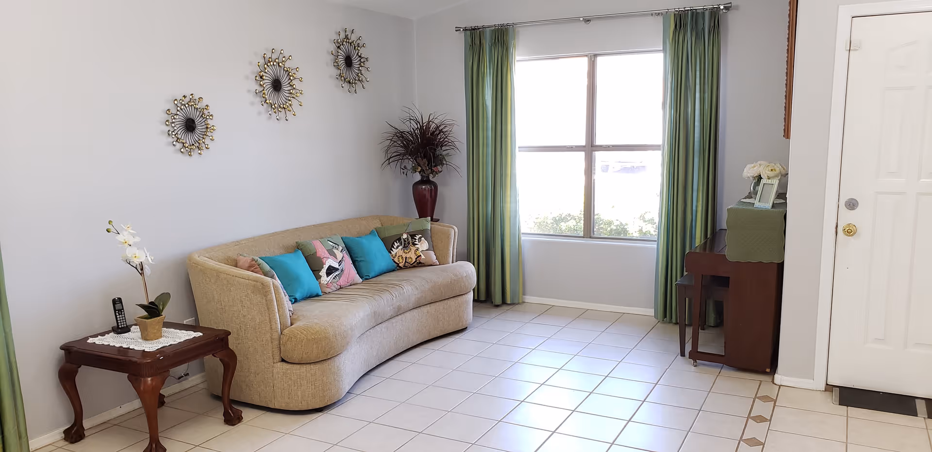Bright living room with a curved beige sofa topped with colorful pillows, a side table, large window with green curtains, and tiled floor.