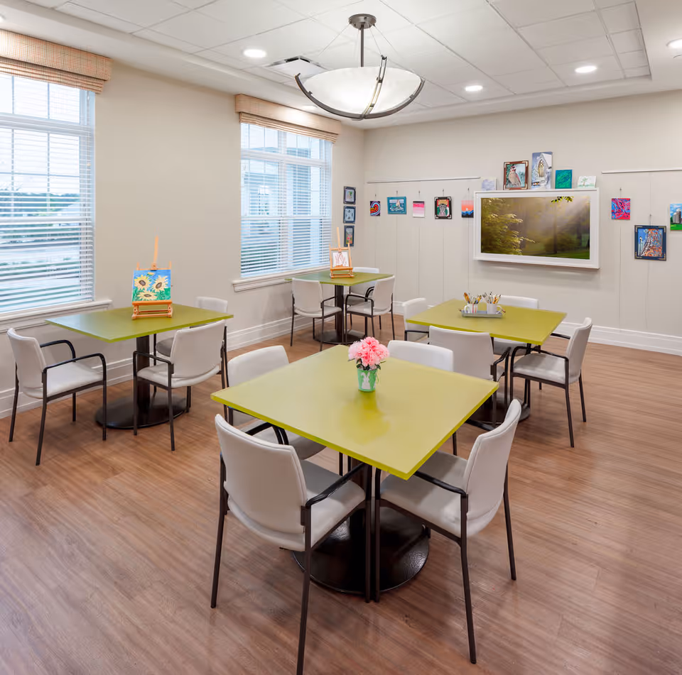 A bright and clean activity room with four green square tables, each surrounded by white chairs. The room has large windows with blinds, wooden flooring, and a ceiling light fixture. Various colorful paintings and artworks are displayed on the walls, and art supplies are placed on the tables.