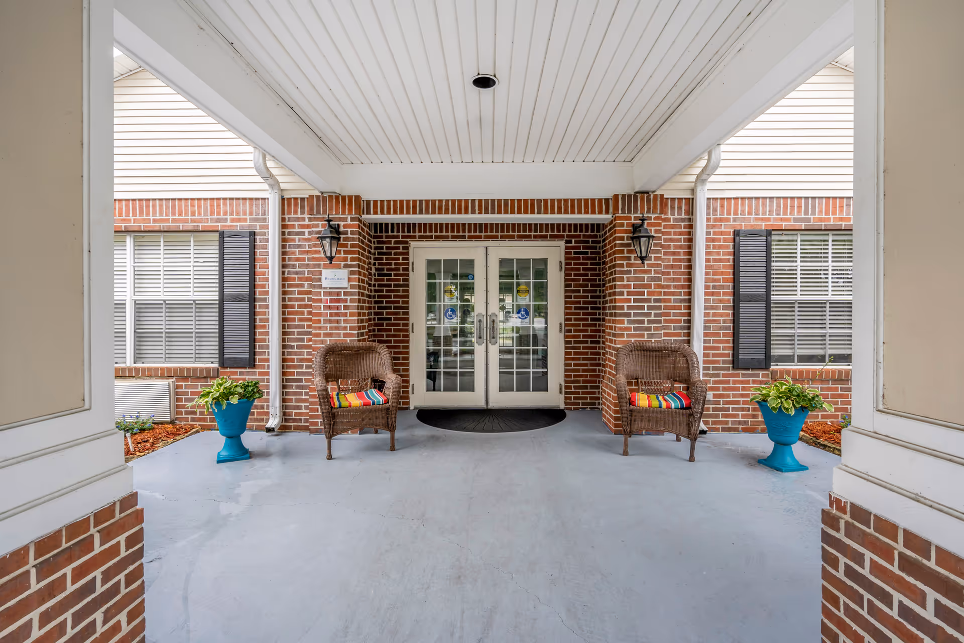 Covered brick entrance with double glass doors, two wicker chairs with colorful cushions, and potted plants.