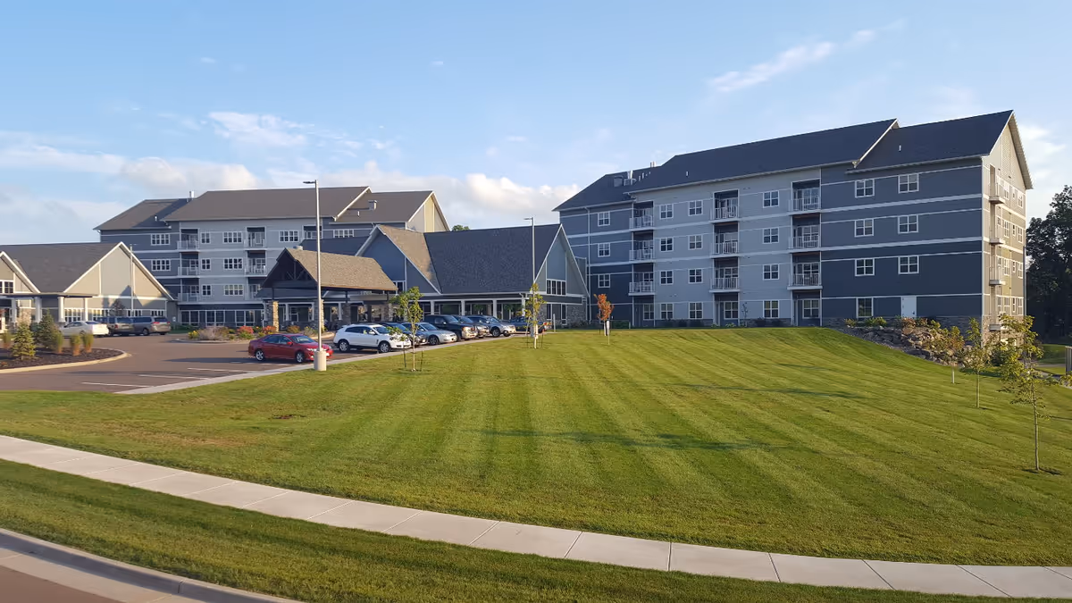 Exterior view of The Classic at Hillcrest Greens, showing multiple connected buildings with gray siding and dark roofs, a parking lot with several cars, a well-maintained green lawn, and a sidewalk in the foreground under a partly cloudy sky.