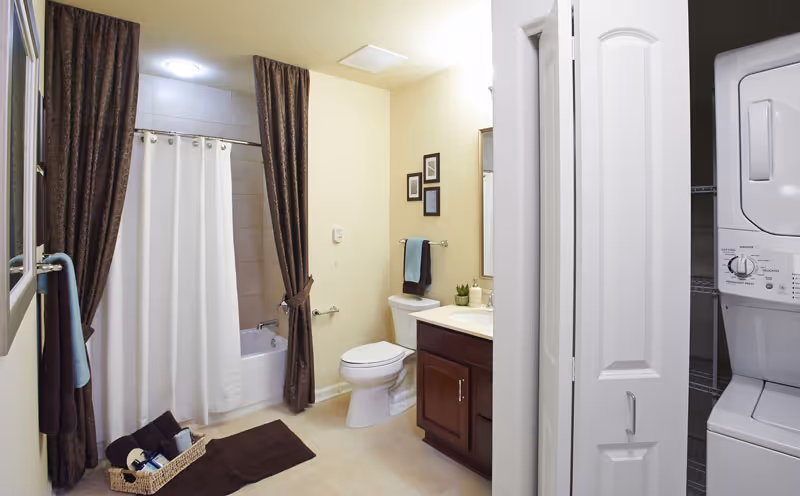 Well-lit bathroom featuring a bathtub/shower with a white curtain, toilet, vanity, decorative dark curtains, and a stacked washer/dryer in a closet.