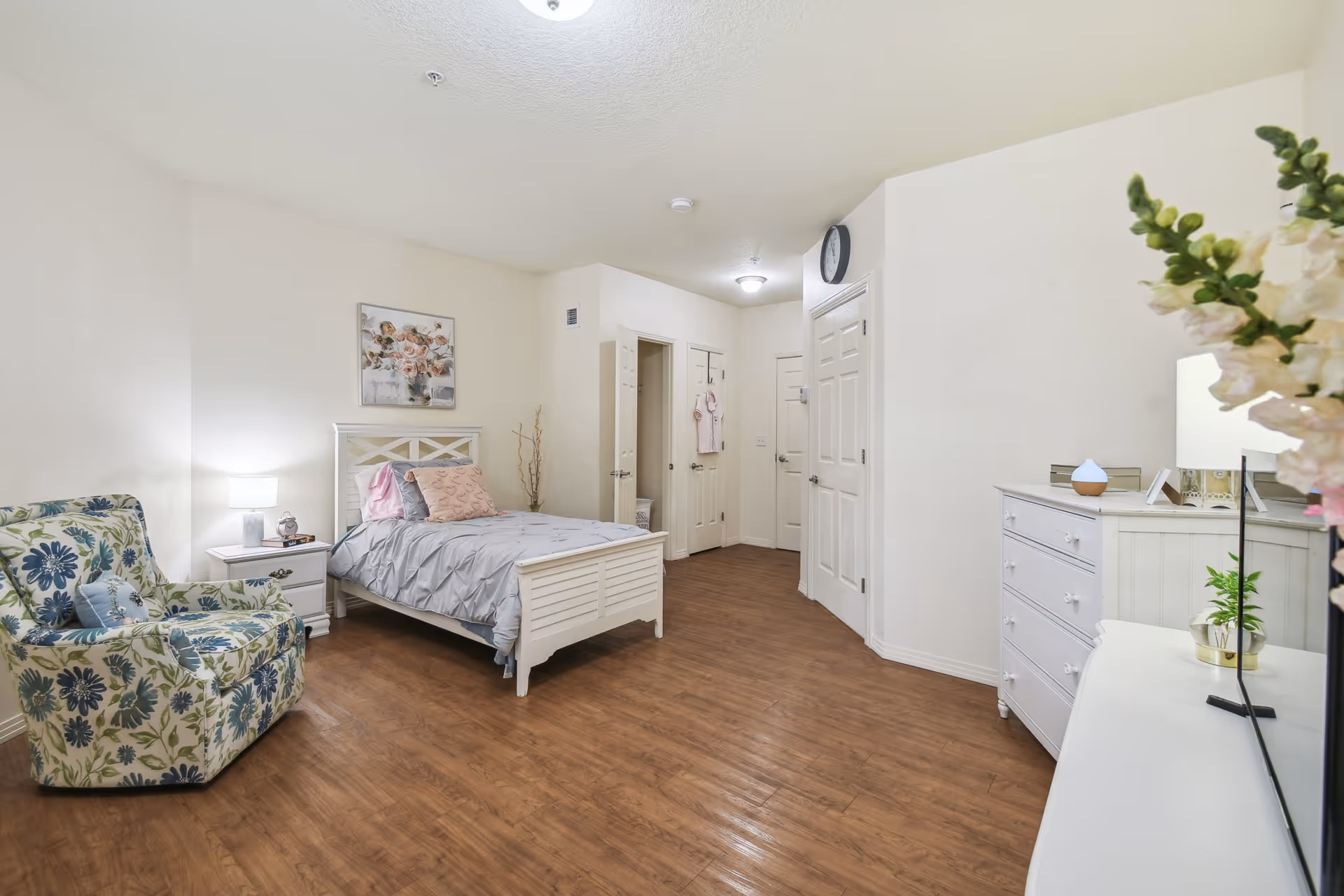 A bright and spacious bedroom with a single bed featuring gray bedding and pink pillows. There is a floral armchair with a blue pillow, a white nightstand with a lamp and clock, a white dresser with a small plant and lamp, and a wall clock above a door. The room has wooden flooring and white walls, with a floral painting hanging above the bed.
