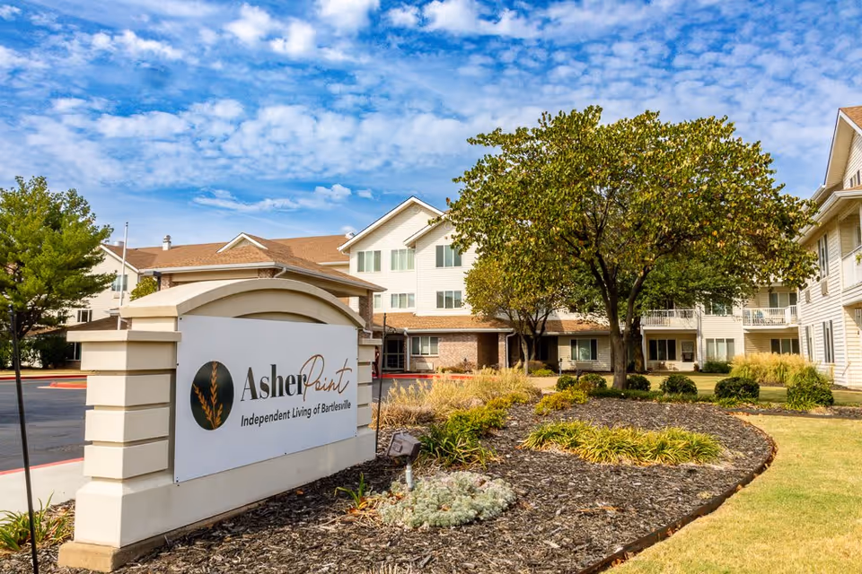 Exterior view of Asher Point Independent Living of Bartlesville facility with a large sign in the foreground, landscaped garden beds, trees, and a multi-story building under a partly cloudy blue sky.