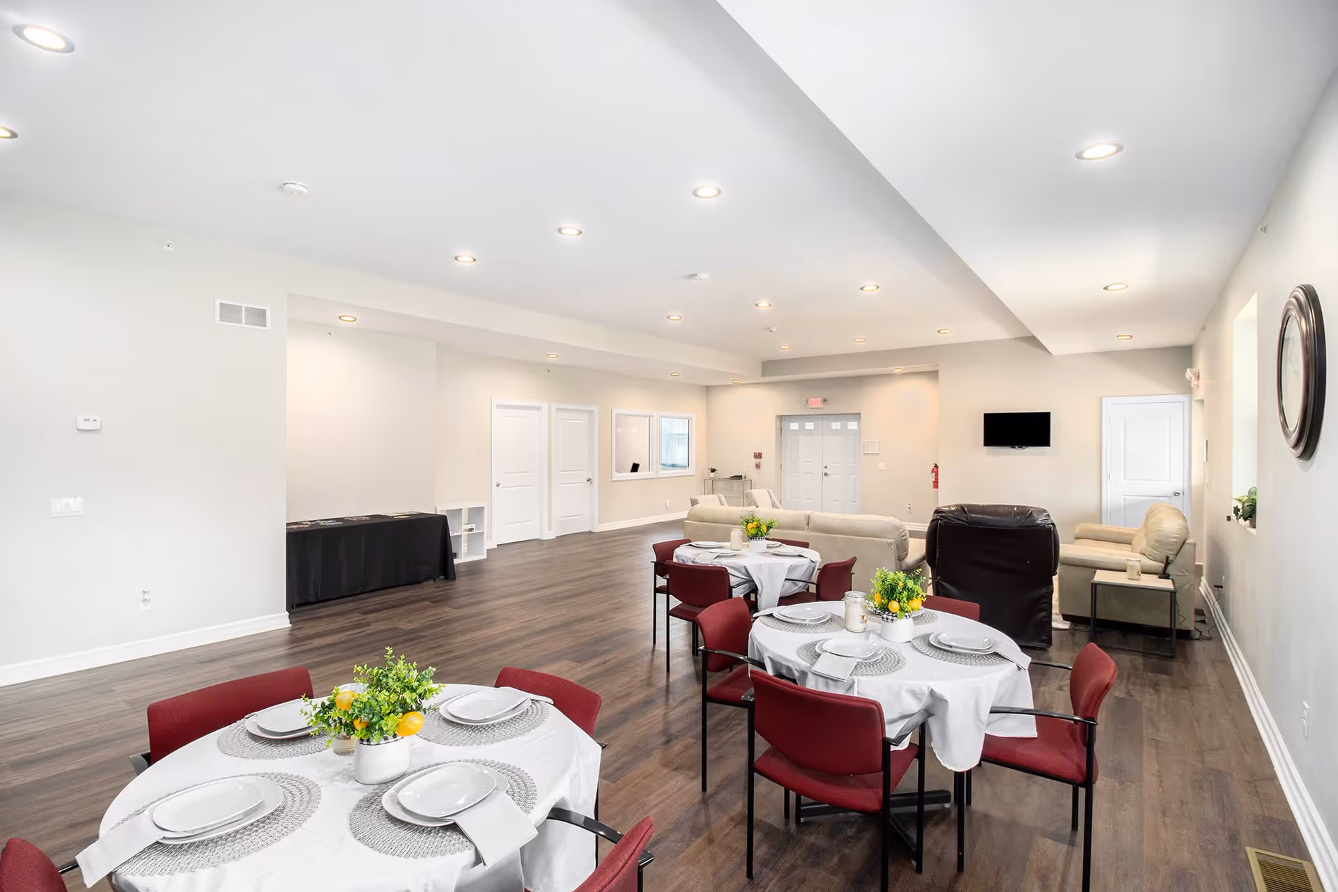 Bright communal dining and lounge room with round tables set for meals and sofas near a wall-mounted TV.