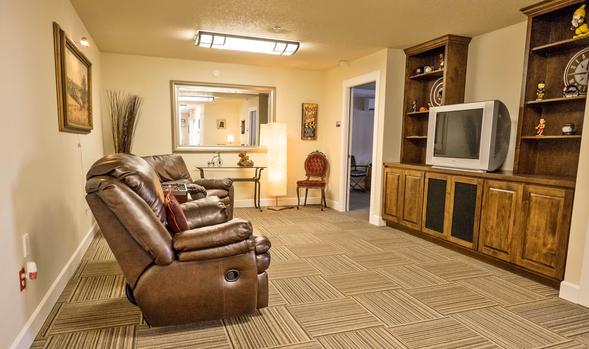 Communal living room with brown leather recliners facing a television in a wooden built-in cabinet, with mirror, lamp, and accent chair in the background.