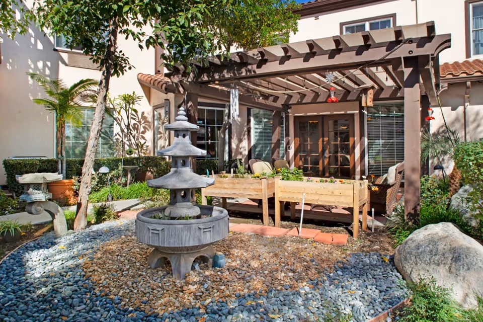 Outdoor garden area at a senior living facility featuring a stone pagoda-style fountain surrounded by small rocks, raised wooden planter boxes with plants, a wooden pergola casting shadows, patio chairs, and various greenery including trees and shrubs near a building with windows and French doors.