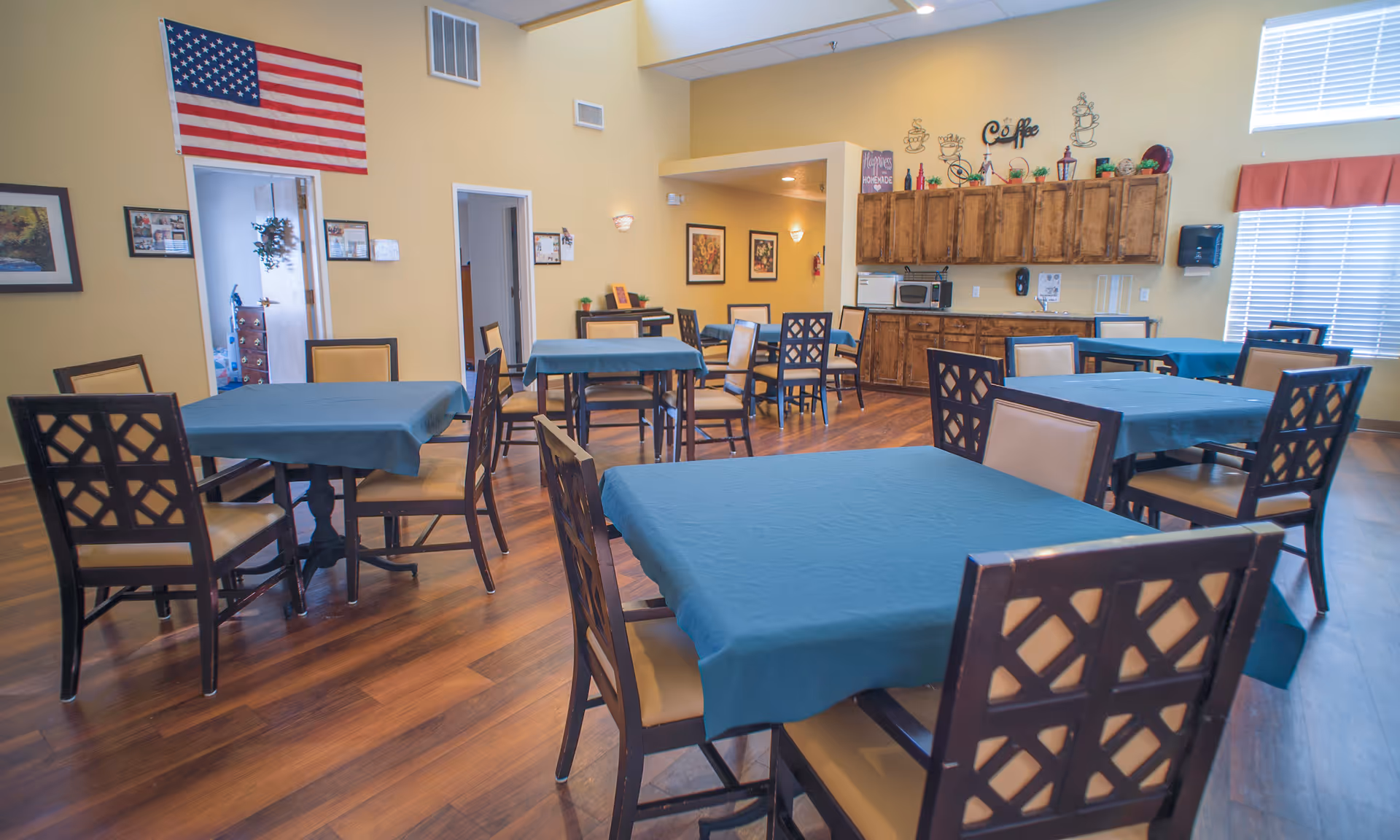 A dining room with several tables covered in blue tablecloths and surrounded by wooden chairs with beige cushions. The room has wooden flooring, yellow walls, and a large American flag hanging on one wall. There is a kitchenette area with wooden cabinets, a microwave, and a coffee sign above. Windows with blinds and a red valance let in natural light.