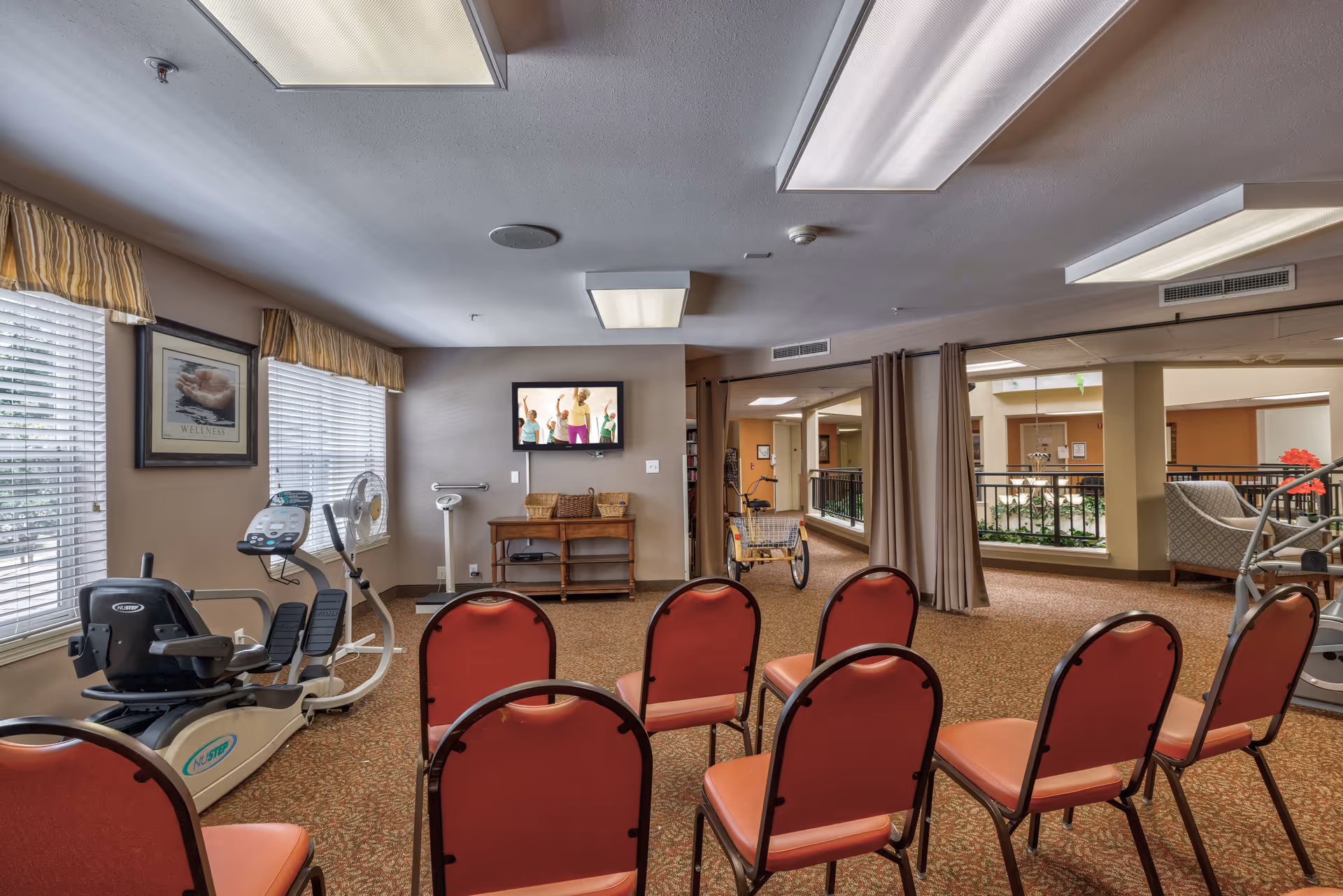 A senior living facility exercise room with rows of red chairs facing a wall-mounted TV showing a fitness class. Exercise equipment is positioned near large windows with blinds and valances. The room has carpeted floors, overhead fluorescent lighting, and a small wooden table with baskets beneath the TV. The space opens into a hallway with additional seating and plants visible.