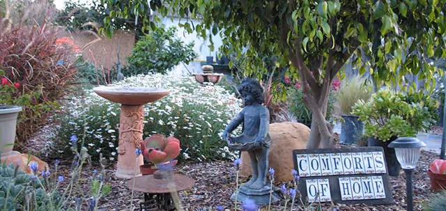 A garden area with various plants and flowers, featuring a birdbath, a small statue of a child holding a basket, and a sign that reads 'COMFORTS OF HOME'.