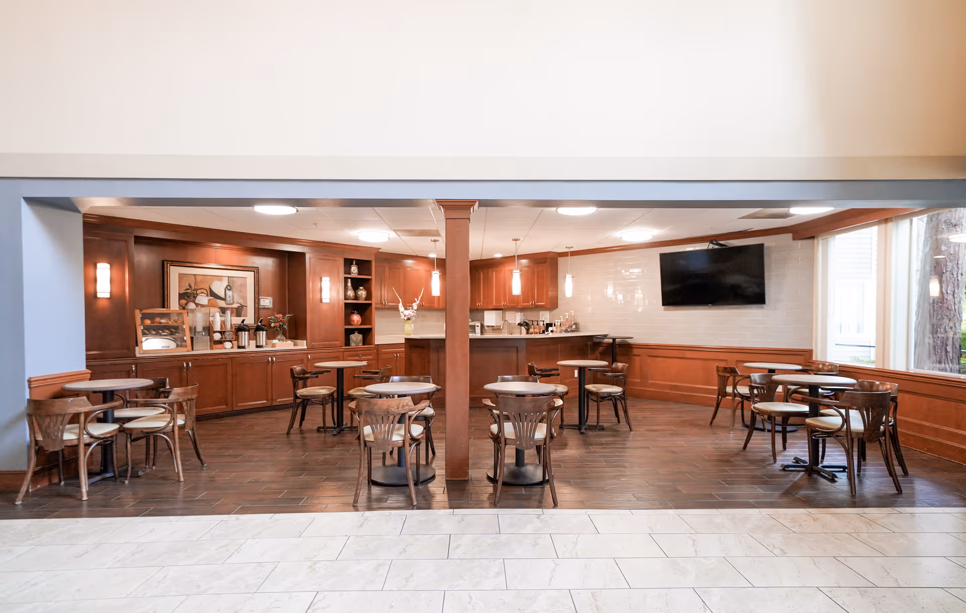 A dining area in an assisted living facility with several round tables and wooden chairs arranged neatly. The room features wood-paneled walls, a counter with coffee and beverage supplies, a large flat-screen TV mounted on the wall, and a large window letting in natural light.