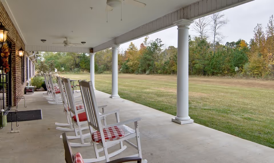 Covered outdoor patio area with white rocking chairs featuring red and white checkered cushions, ceiling fans, and white columns overlooking a grassy lawn and trees in the background.