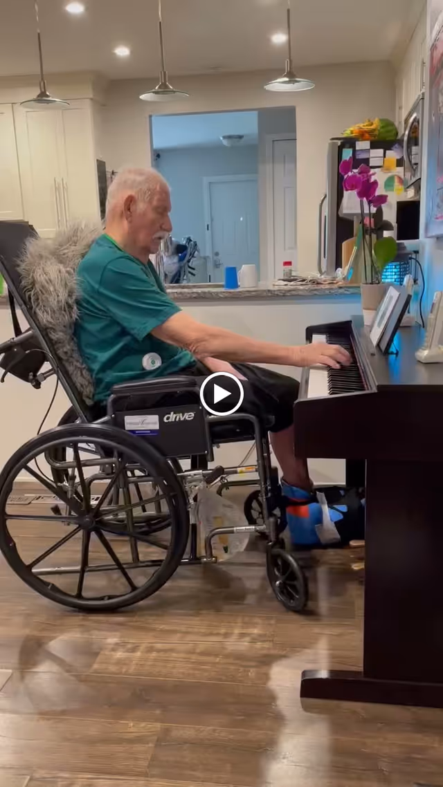 An elderly man in a wheelchair playing a piano in a well-lit room with wooden flooring. The room has a kitchen counter in the background with hanging lights above it, and a vase with purple flowers is placed on the piano.