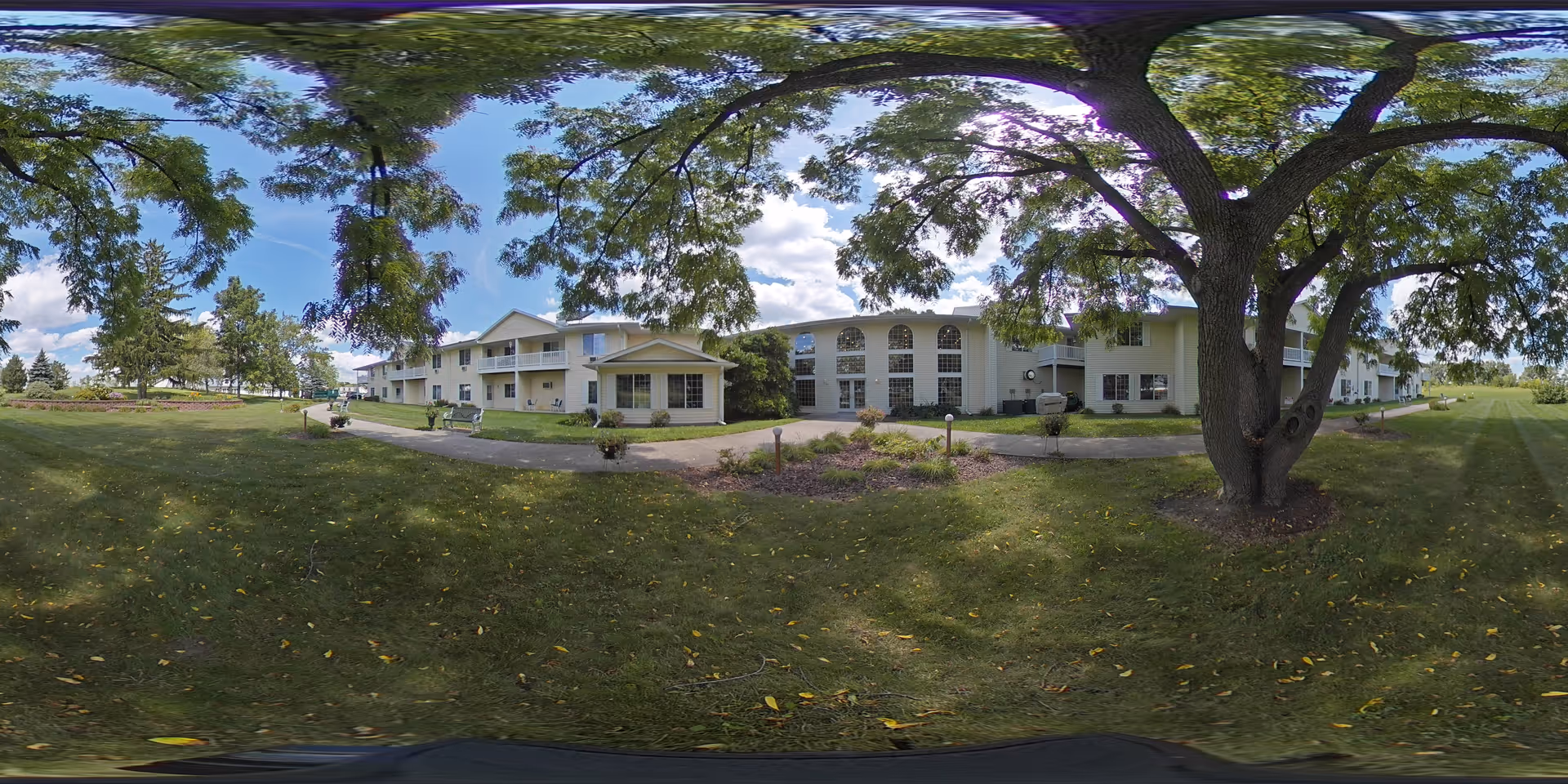Panoramic view of a two-story assisted living building with a large front lawn, walking paths, benches, and mature shade trees.