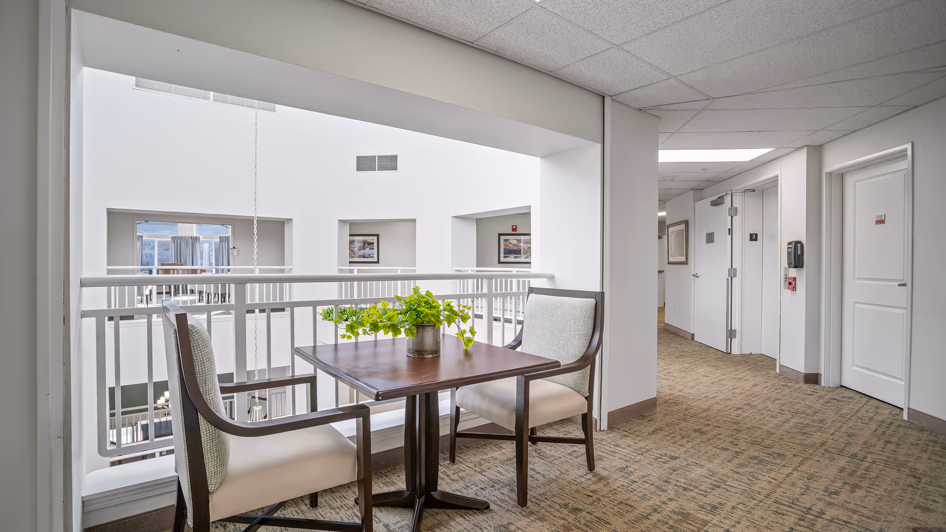 A small seating area with a wooden table and two cushioned chairs placed near a railing overlooking a bright atrium with white walls and framed pictures. The floor is carpeted and there is a hallway with several white doors and a hand sanitizer dispenser on the wall.