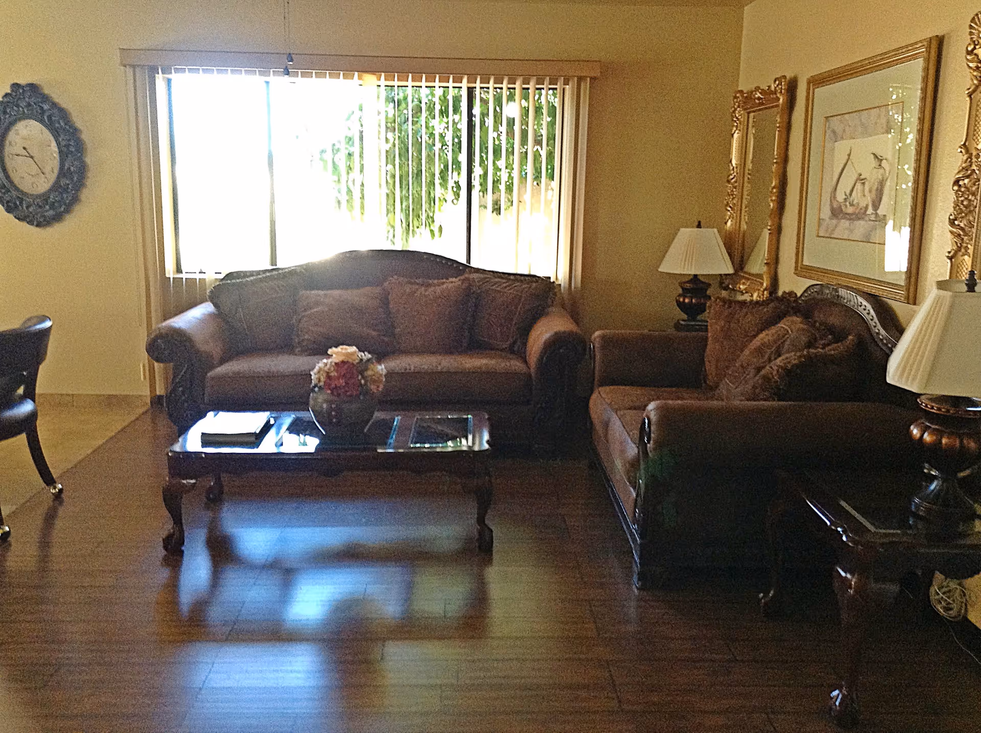A living room with two brown upholstered sofas, a wooden coffee table with a flower vase on it, two side tables each with a lamp, a wall clock, and framed artwork on the walls. There is a large window with vertical blinds letting in natural light.