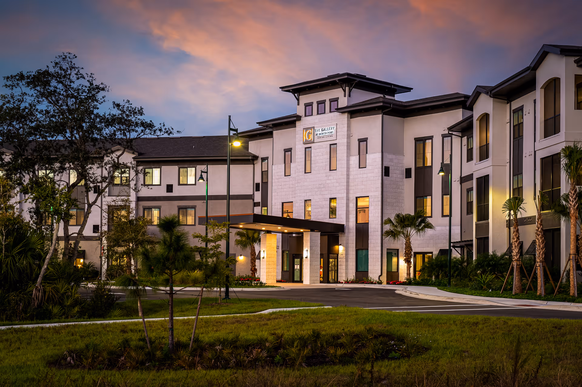 Exterior view of a modern senior living facility building at dusk with warm interior lights visible through the windows, surrounded by landscaped greenery and palm trees under a colorful sky.