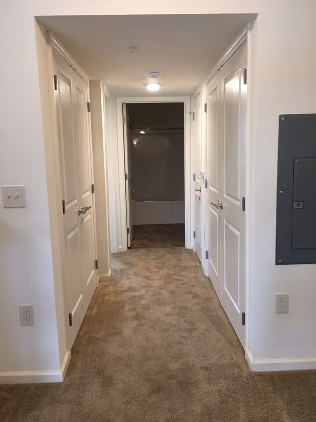 A carpeted hallway with white walls and multiple closed white doors on both sides. At the end of the hallway, there is an open door revealing a bathroom with a bathtub and tiled floor. A gray electrical panel is mounted on the right wall.
