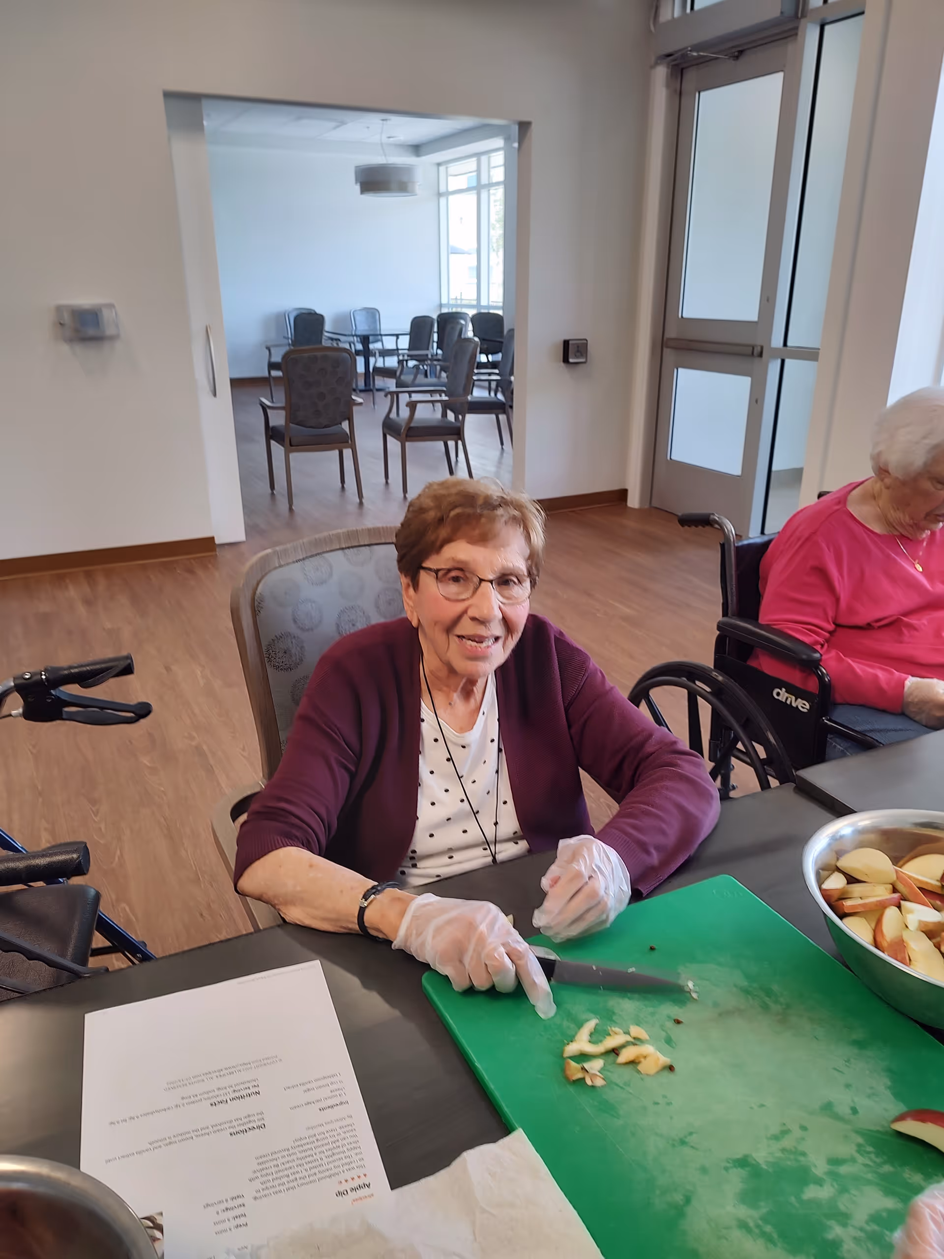 An elderly woman wearing gloves smiles while cutting apples on a green cutting board in a senior living common room.