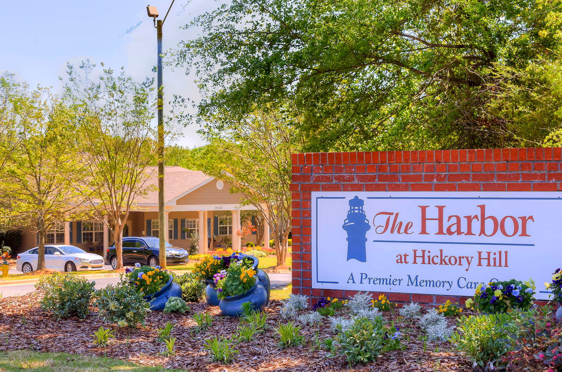 Exterior view of The Harbor at Hickory Hill facility with a brick sign displaying the name and description as a premier memory care center. The foreground has landscaped flower beds with colorful flowers and greenery. In the background, there is a building with a covered entrance and several parked cars, surrounded by trees and greenery.