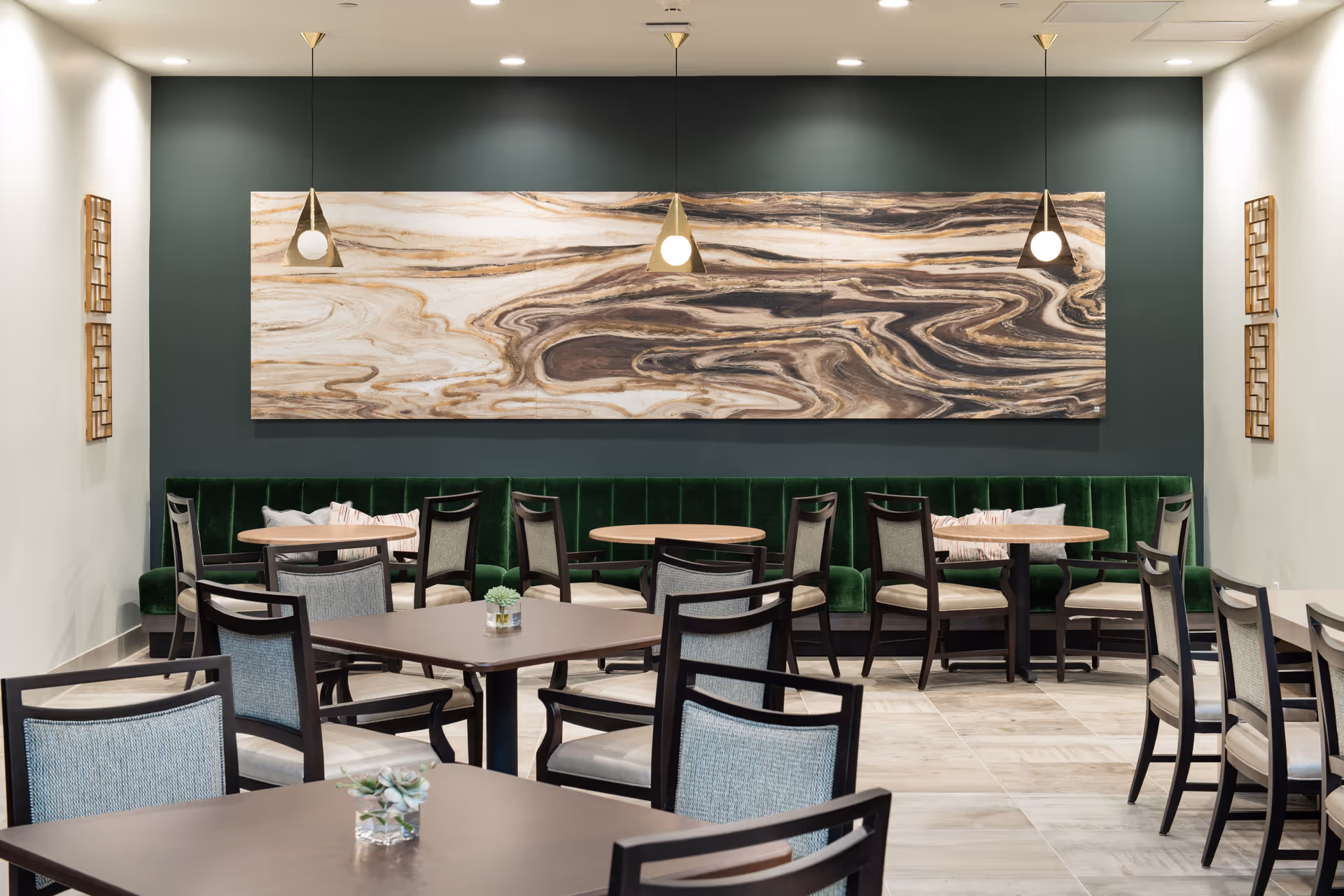 A modern dining area with multiple tables and chairs arranged neatly. The back wall features a large abstract wood-patterned artwork above a green velvet bench. Three pendant lights hang from the ceiling, illuminating the space. The room has light-colored tiled flooring and neutral walls with decorative wall accents.