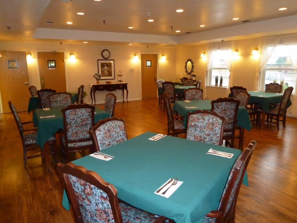 Dining room with multiple tables covered in green tablecloths and floral-upholstered chairs on a hardwood floor.
