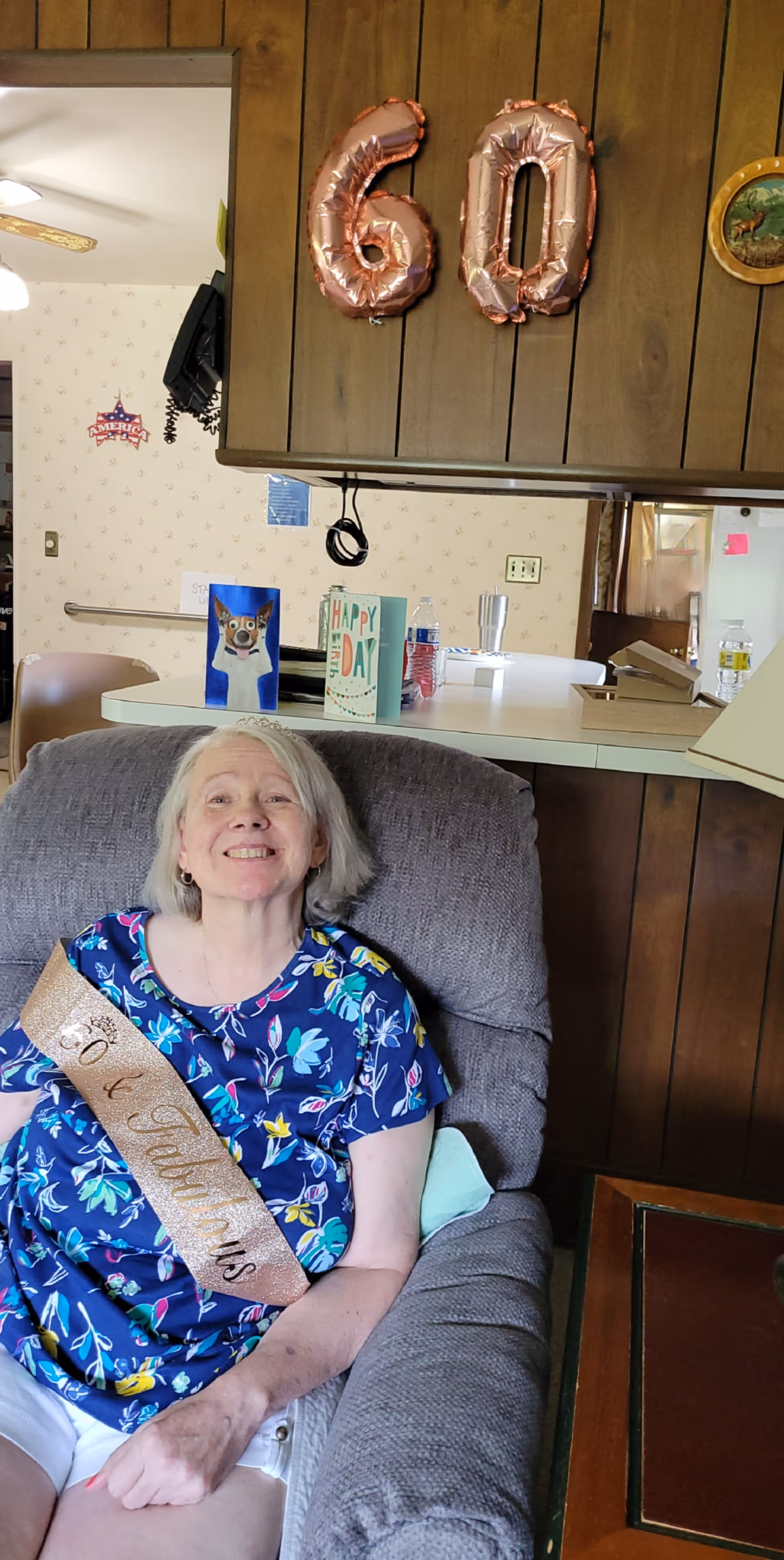 An elderly woman with gray hair wearing a blue floral shirt and white shorts sits smiling in a gray armchair. She is wearing a gold sash that reads '60 & Fabulous'. Behind her, rose gold balloons shaped as the number 60 are attached to a wooden wall. On the counter behind her, there are birthday cards and some cups. The setting appears to be a cozy living room with wood paneling and a kitchen area visible in the background.