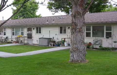Single-story senior living building with green lawn, walkway, and large tree in foreground.