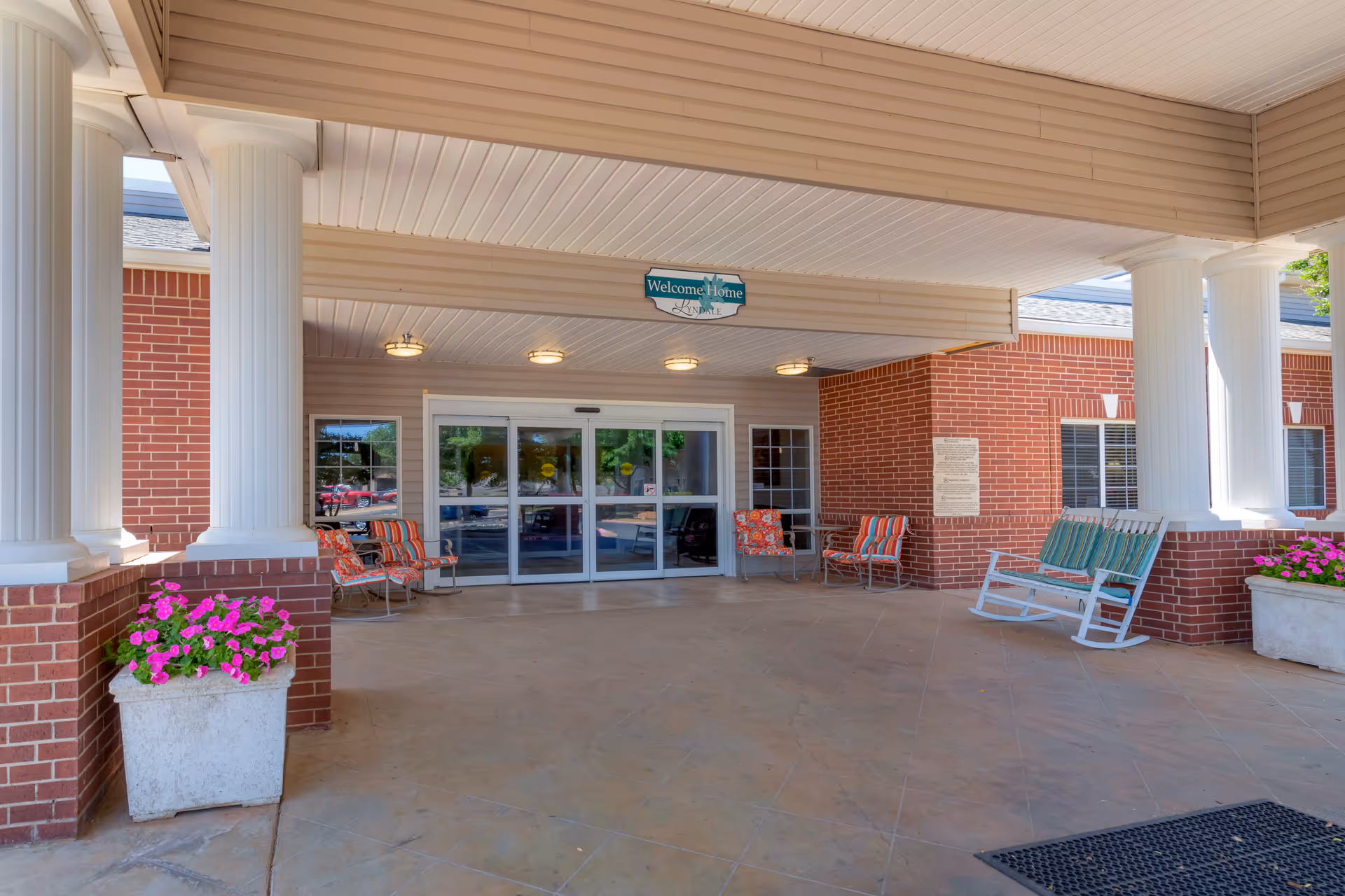Entrance area of a senior living facility with large white columns, brick walls, and a covered drop-off area. There are several chairs with colorful cushions and a white rocking chair arranged near the automatic glass doors. Two large planters with pink flowers are placed on either side of the entrance. A sign above the doors reads 'Welcome Home'.