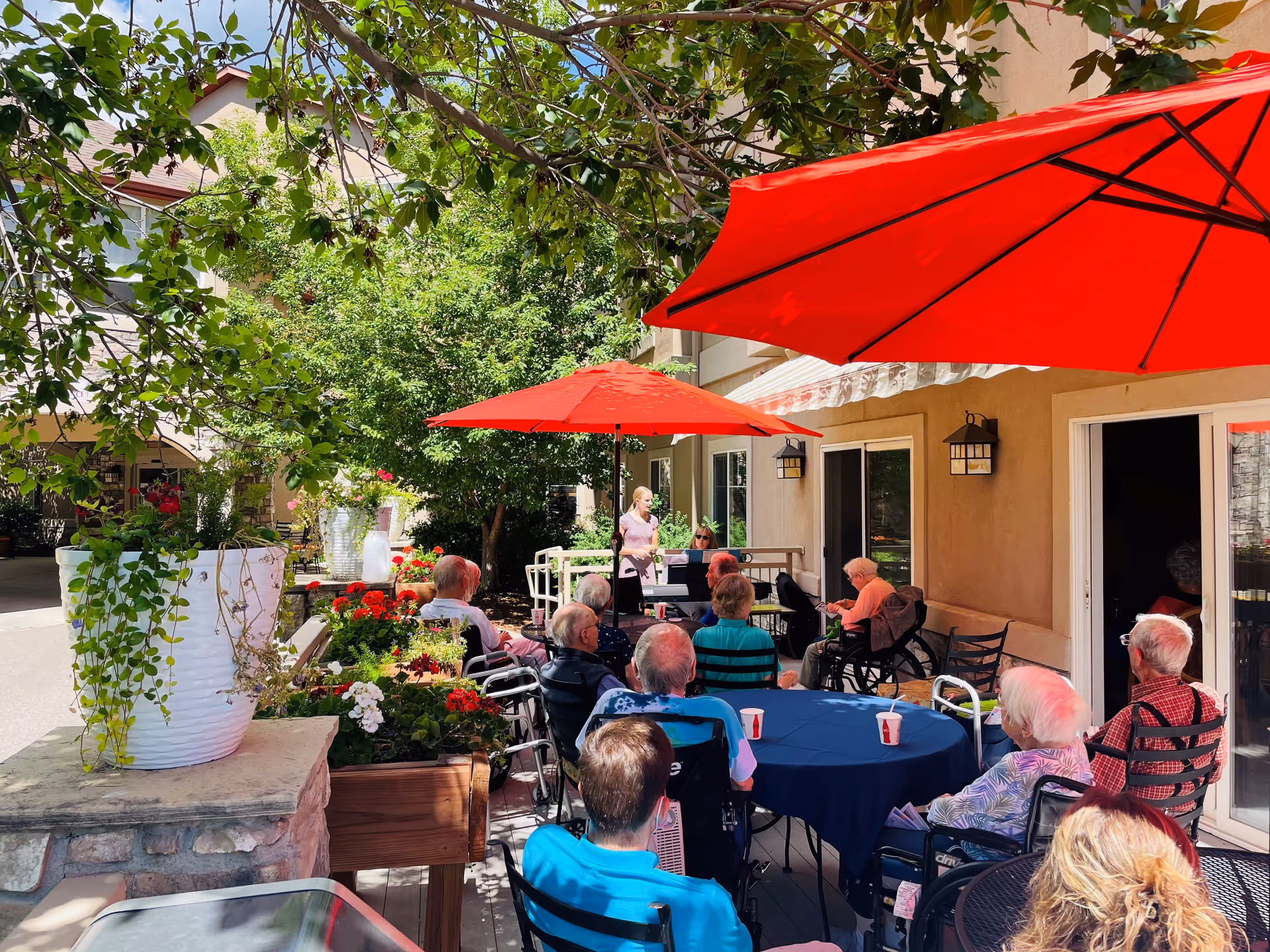 Residents seated at tables under red umbrellas on an outdoor patio listening to a performer by the building.