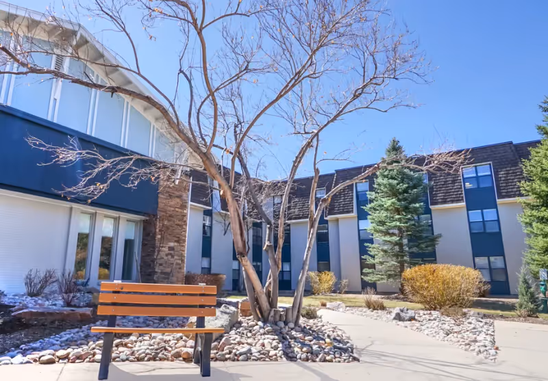 Outdoor courtyard area of a senior living facility with a wooden bench, a leafless tree surrounded by rocks, and a pathway. The building in the background has multiple windows and a mix of stone and siding exterior under a clear blue sky.