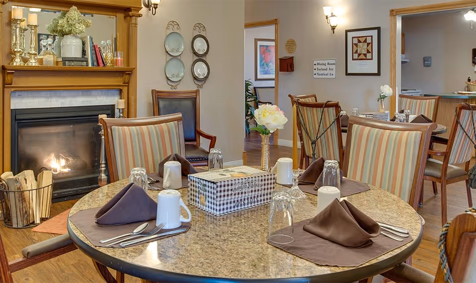 A cozy dining area in a senior living facility with a round table set for four, featuring brown napkins, white mugs, and upside-down glasses. The room has striped cushioned chairs, a lit fireplace with decorative items on the mantle, and framed artwork on the walls.