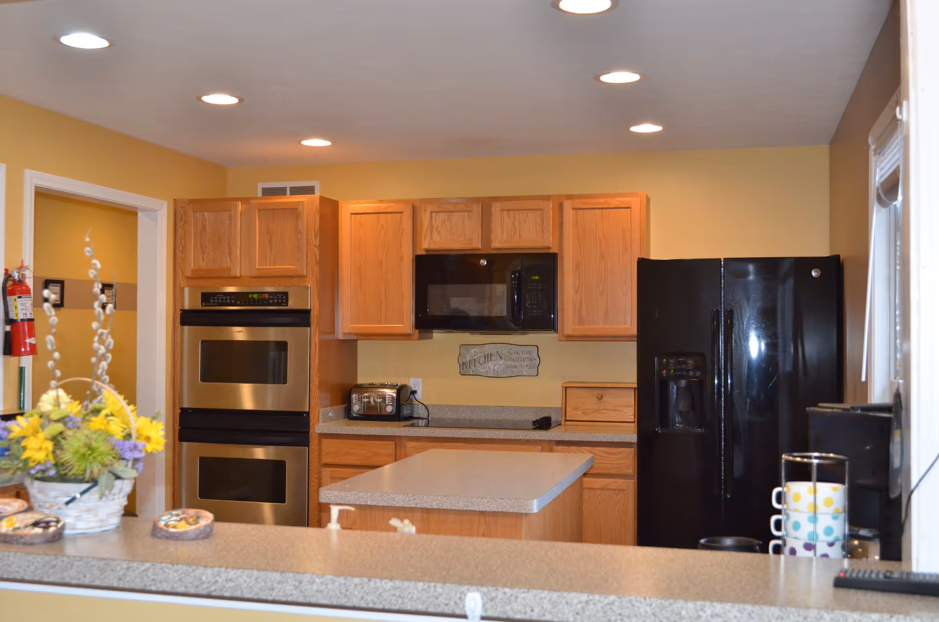 Interior view of a kitchen with wooden cabinets, a black refrigerator, a black microwave, a double oven, a toaster, and a kitchen island. There is a basket with yellow and purple flowers on the counter in the foreground.
