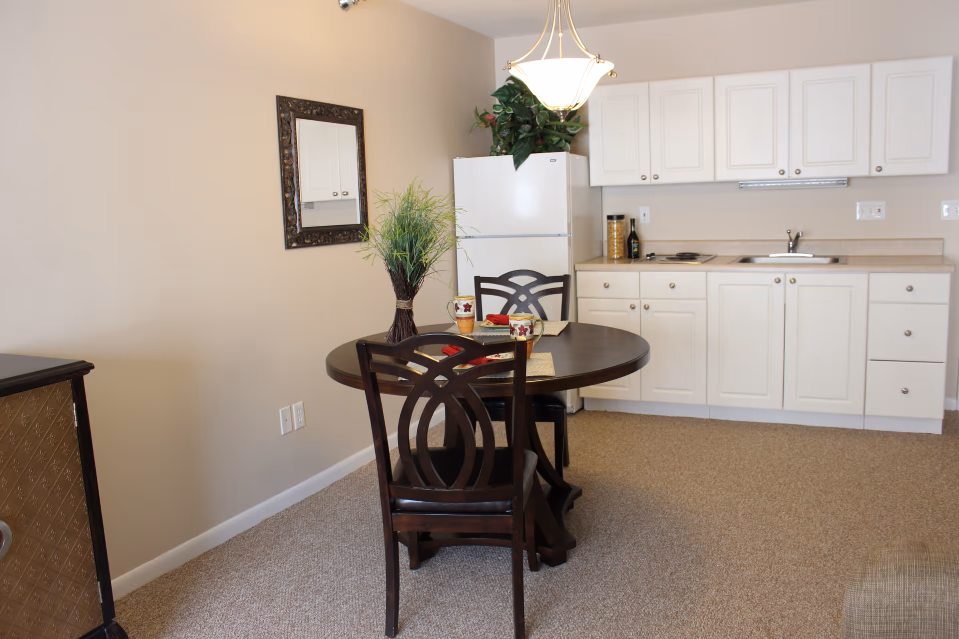 A small kitchen and dining area with white cabinets, a white refrigerator, a sink, and a stove. In front of the kitchen is a round dark wooden dining table with two matching chairs. The table has a plant centerpiece and two mugs on placemats. A decorative mirror hangs on the beige wall, and a hanging light fixture is above the table.
