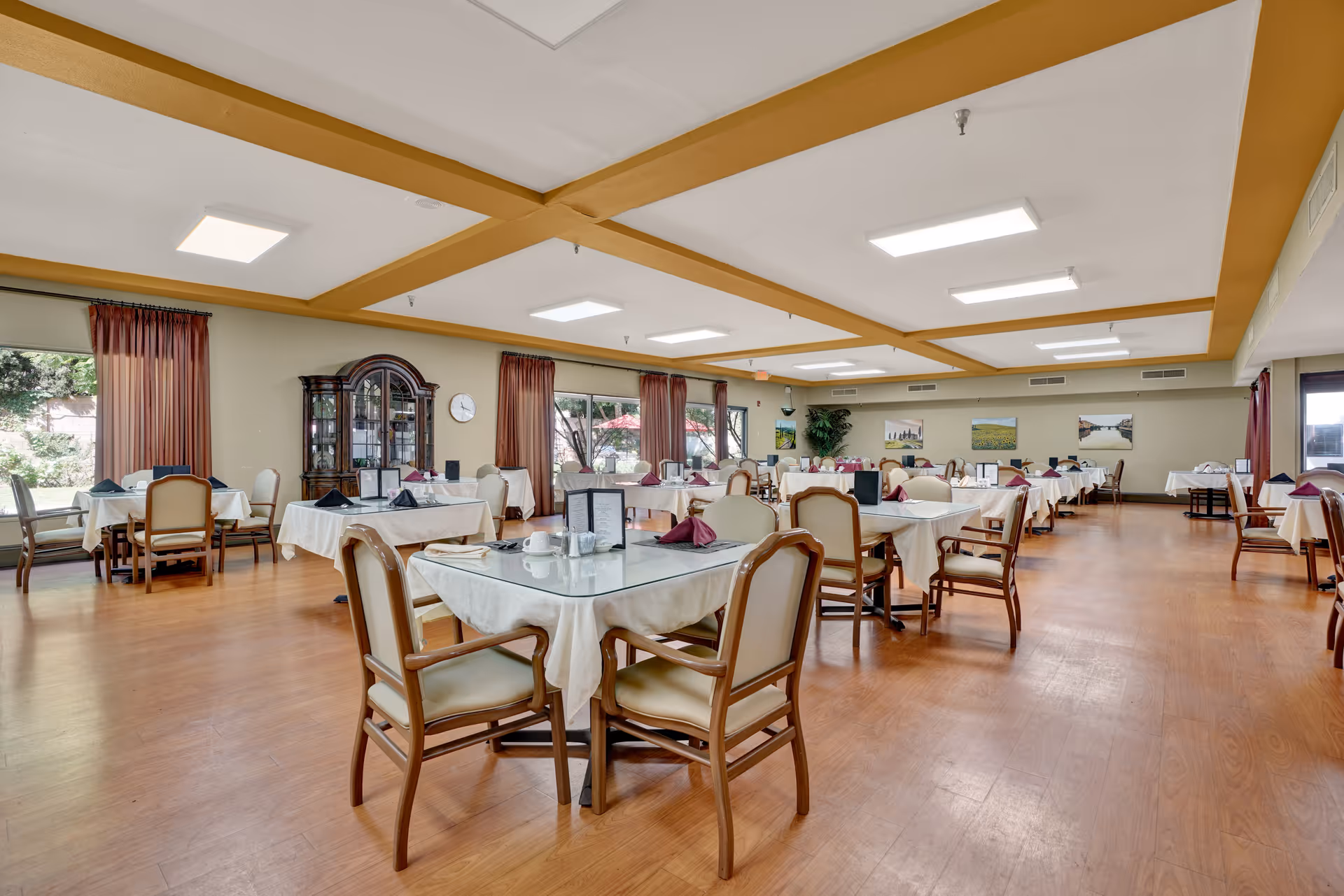 Spacious dining room with multiple tables covered in white tablecloths and set with napkins and cups. The room has wooden flooring, beige walls, large windows with red curtains, and ceiling lights. There is a wooden cabinet and some framed pictures on the walls.