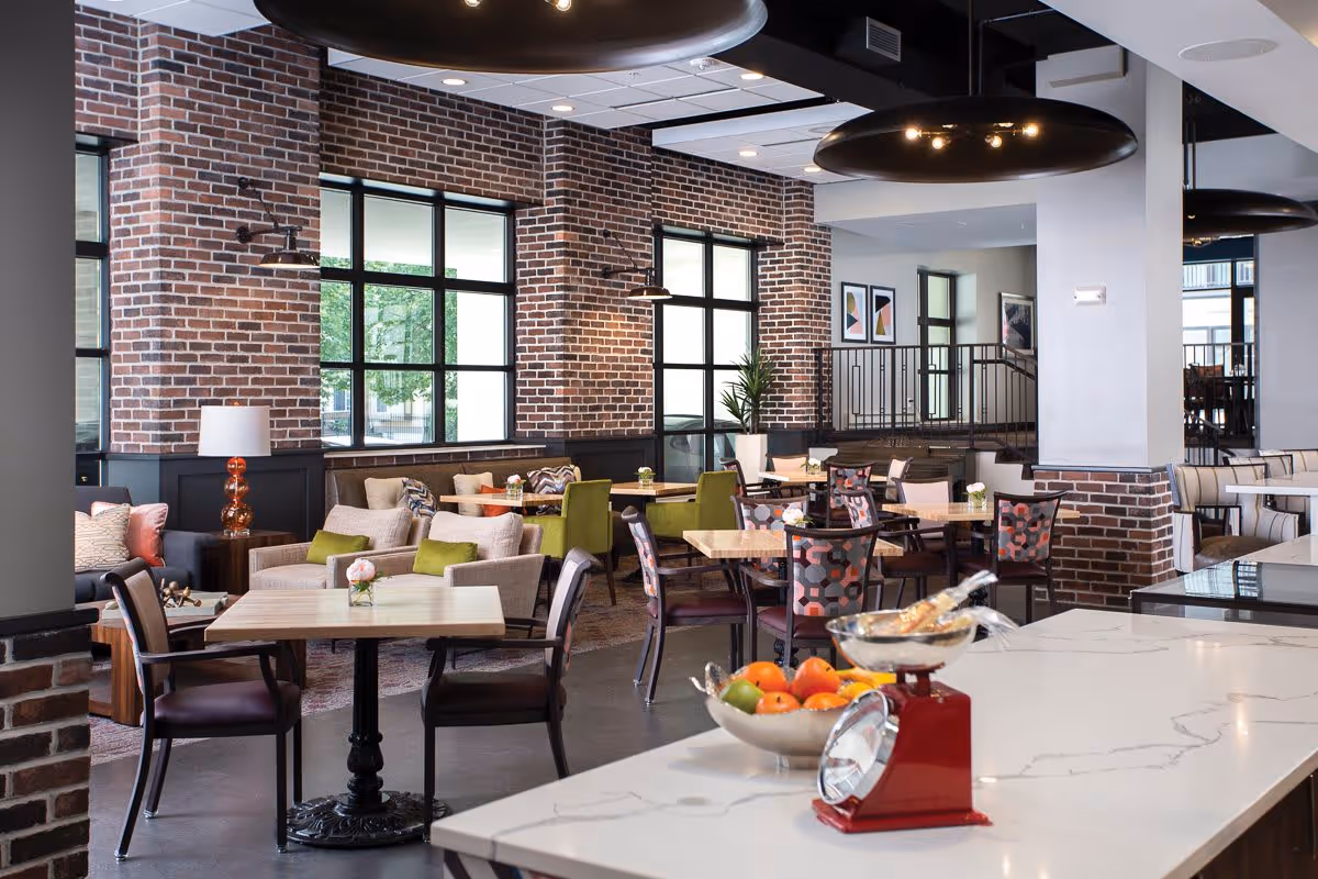 A spacious and modern dining area with brick walls and large windows letting in natural light. The room features various seating options including tables with chairs and cushioned armchairs, some with green and patterned upholstery. A white marble countertop in the foreground holds a bowl of fresh fruit and a red kitchen scale. The ceiling has recessed lighting and large black circular light fixtures.