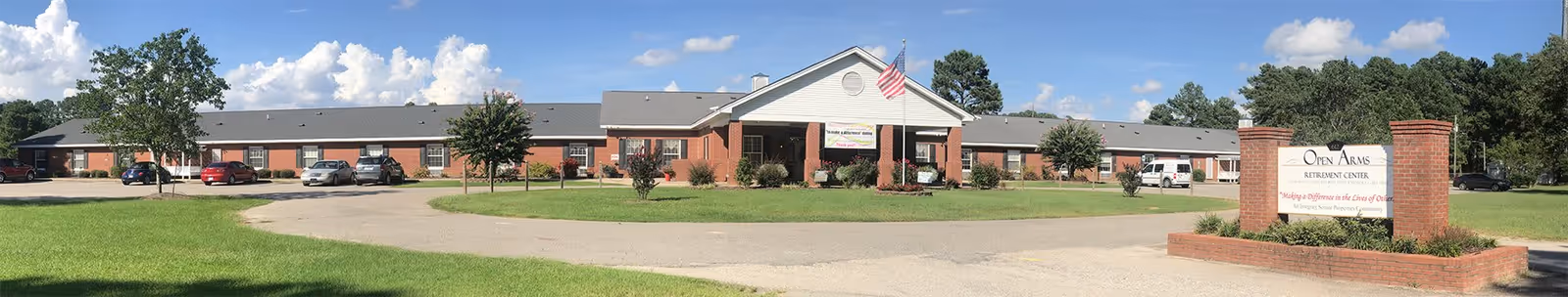 Front exterior view of Open Arms Retirement Center, a single-story brick building with a gray roof, an American flag on a pole in front, several parked cars, and a sign displaying the facility's name and slogan on a brick base. The sky is blue with scattered clouds and there is green grass and trees around the building.