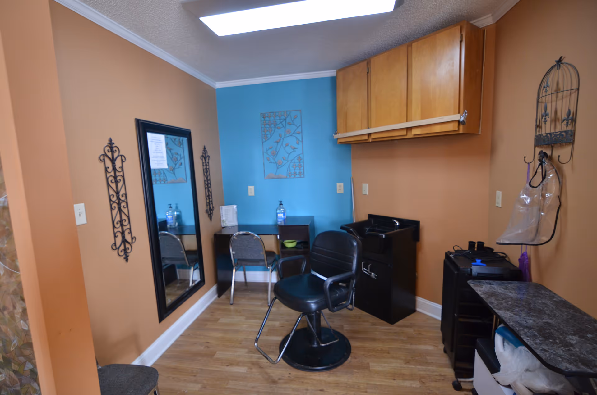 A small salon room with a black salon chair in the center, a black sink station, a desk with a chair against a blue accent wall, and wooden cabinets mounted on the wall. The room has light brown walls, a large mirror on one wall, decorative metal wall hangings, and a wooden floor.